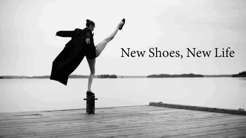 Bilden är i svartvitt och visar en person som står på en brygga vid vattnet. Personen balanserar elegant på en stolpe med ena benet högt uppsträckt i luften, vilket ger en känsla av rörelse och balans. De bär en lång, mörk rock som fladdrar något i luften, samt skor. Bakgrunden är stillsam med en spegelblank sjö och några öar eller landkonturer i fjärran under en molnig himmel. Bilden förmedlar en känsla av frihet, styrka och konstnärlig uttrycksfullhet, med en tydlig kontrast mellan den dynamiska posen och det lugna landskapet. Innehåller texten New Shoes, New Life.