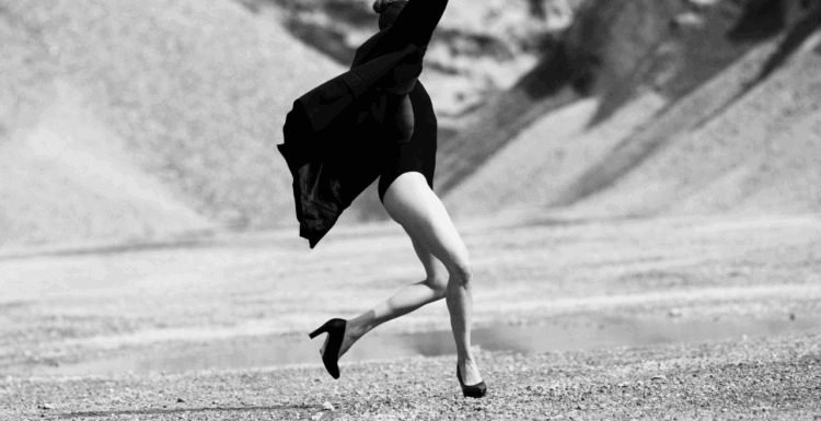 Bilden är i svartvitt och visar en person i rörelse på ett kargt, stenigt landskap med berg eller sanddyner i bakgrunden. Personen verkar mitt i ett språng eller en dansliknande gest, med armarna lyfta och kroppen i dynamisk rörelse. De bär en mörk dräkt eller body, en lång rock som fladdrar bakom, och högklackade skor.