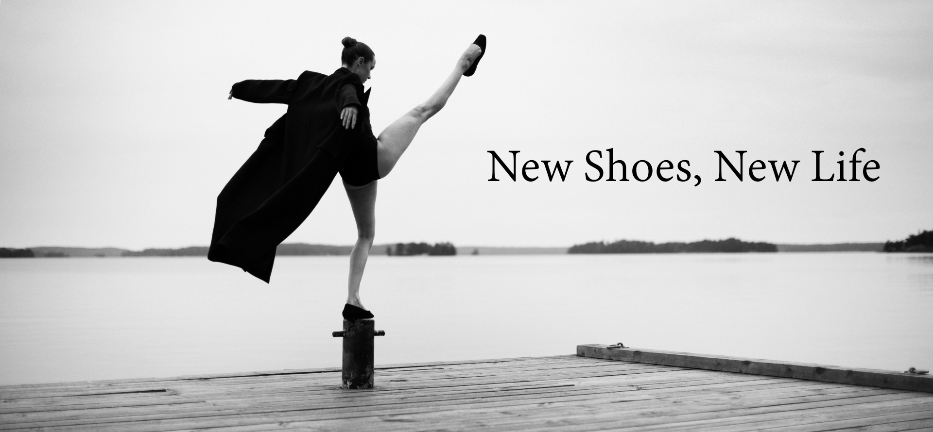 Bilden är i svartvitt och visar en person som står på en brygga vid vattnet. Personen balanserar elegant på en stolpe med ena benet högt uppsträckt i luften, vilket ger en känsla av rörelse och balans. De bär en lång, mörk rock som fladdrar något i luften, samt skor. Bakgrunden är stillsam med en spegelblank sjö och några öar eller landkonturer i fjärran under en molnig himmel. Bilden förmedlar en känsla av frihet, styrka och konstnärlig uttrycksfullhet, med en tydlig kontrast mellan den dynamiska posen och det lugna landskapet. Innehåller texten New Shoes, New Life.