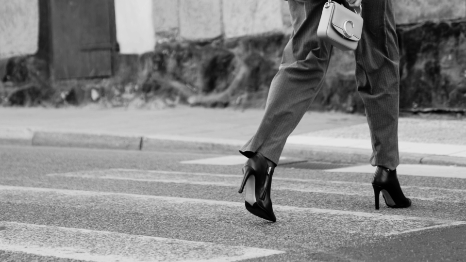 black and white picture of a woman in slacks walking across the street. She is wearing high heel orthopedic boots.