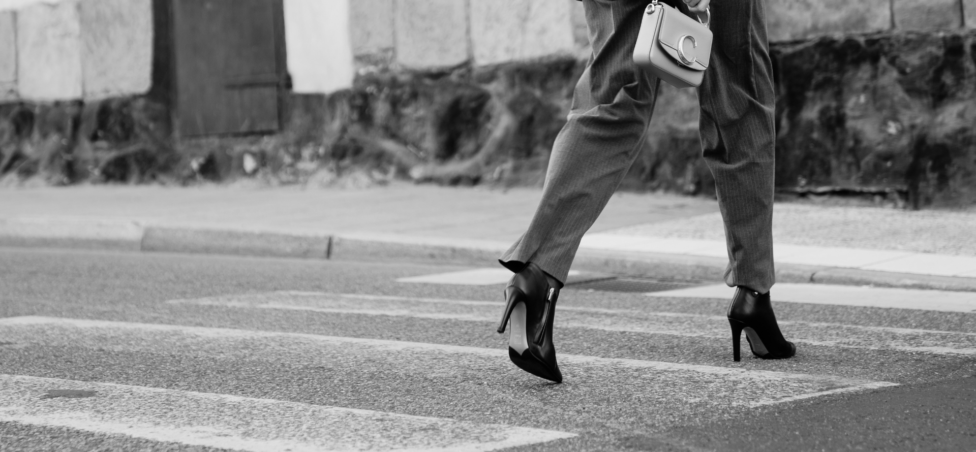 black and white picture of a woman in slacks walking across the street. She is wearing high heel orthopedic boots.