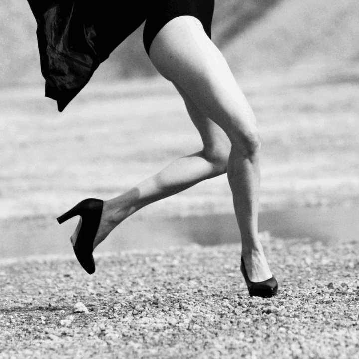Bilden är i svartvitt och visar en person i rörelse på ett kargt, stenigt landskap med berg eller sanddyner i bakgrunden. Personen verkar mitt i ett språng eller en dansliknande gest, med armarna lyfta och kroppen i dynamisk rörelse. De bär en mörk dräkt eller body, en lång rock som fladdrar bakom, och högklackade skor.