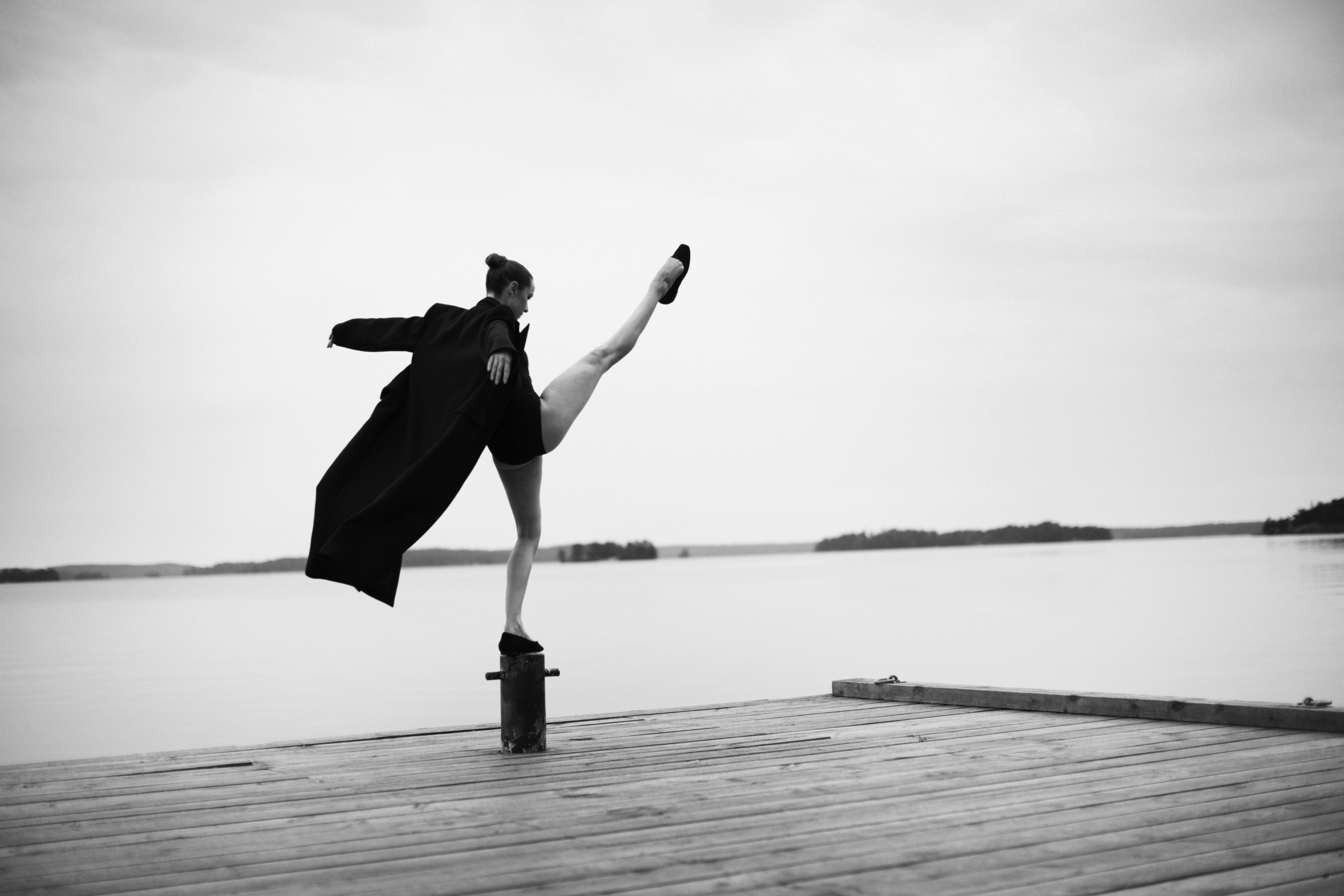 Bilden är i svartvitt och visar en person som står på en brygga vid vattnet. Personen balanserar elegant på en stolpe med ena benet högt uppsträckt i luften, vilket ger en känsla av rörelse och balans. De bär en lång, mörk rock som fladdrar något i luften, samt skor. Bakgrunden är stillsam med en spegelblank sjö och några öar eller landkonturer i fjärran under en molnig himmel. Bilden förmedlar en känsla av frihet, styrka och konstnärlig uttrycksfullhet, med en tydlig kontrast mellan den dynamiska posen och det lugna landskapet.