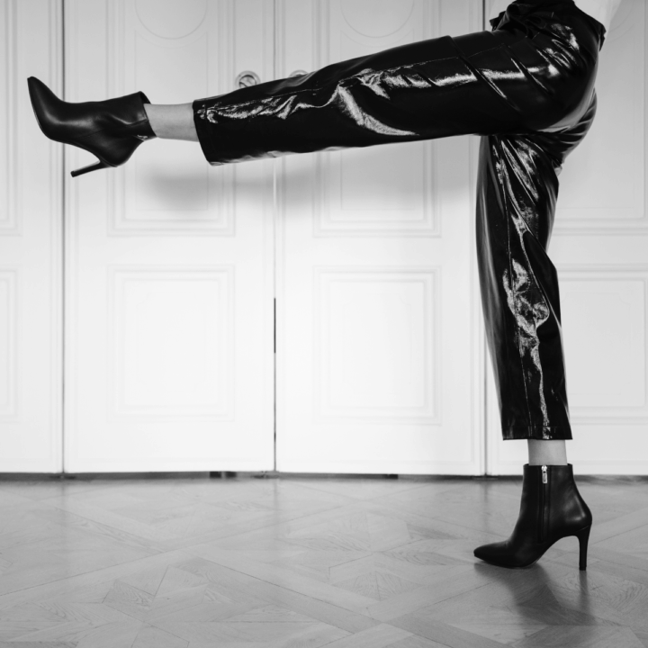 En person mot vit vägg. Personen har svarta glansiga byxor och står i med ena benet utsträckt. På fötterna har hen svarta ortopediska stövletter.