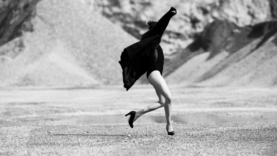 Bilden är i svartvitt och visar en person i rörelse på ett kargt, stenigt landskap med berg eller sanddyner i bakgrunden. Personen verkar mitt i ett språng eller en dansliknande gest, med armarna lyfta och kroppen i dynamisk rörelse. De bär en mörk dräkt eller body, en lång rock som fladdrar bakom, och högklackade skor.