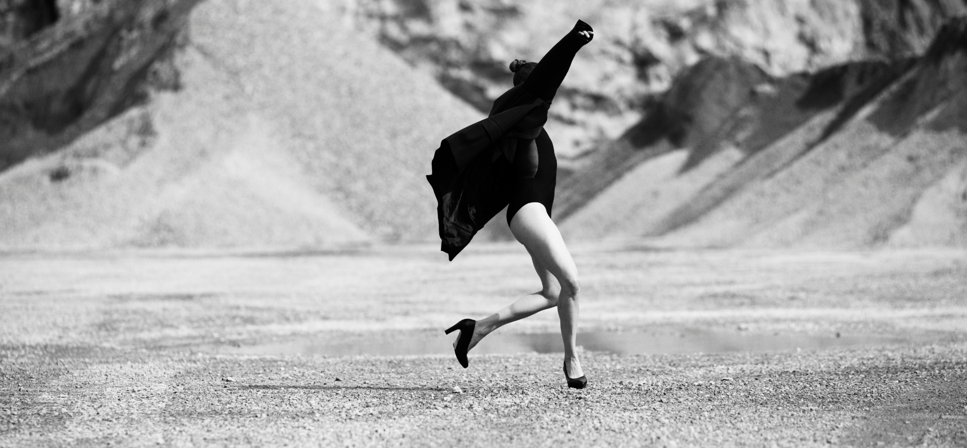 Bilden är i svartvitt och visar en person i rörelse på ett kargt, stenigt landskap med berg eller sanddyner i bakgrunden. Personen verkar mitt i ett språng eller en dansliknande gest, med armarna lyfta och kroppen i dynamisk rörelse. De bär en mörk dräkt eller body, en lång rock som fladdrar bakom, och högklackade skor.