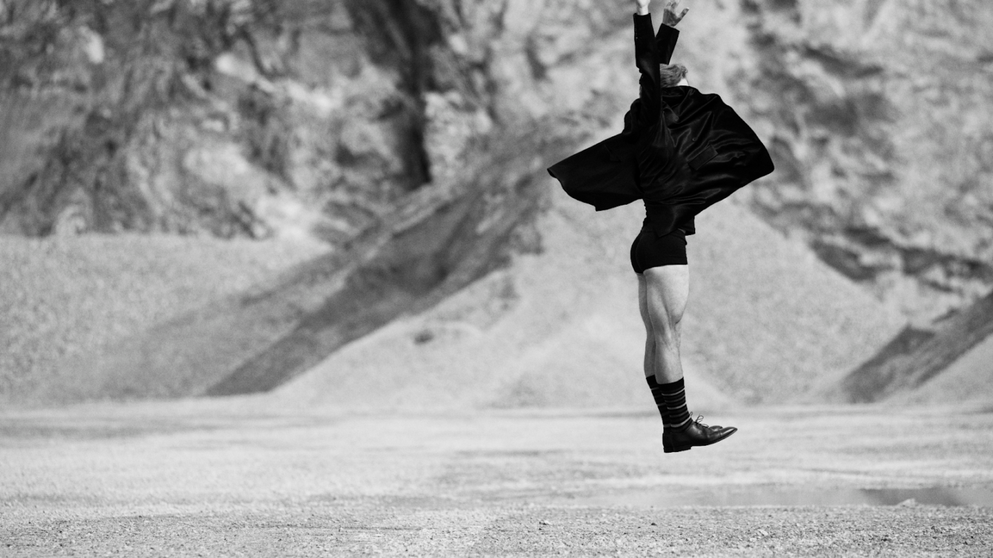 Bilden är i svartvitt och visar en person som hoppar eller svävar i luften i ett kargt, stenigt landskap – kanske en öken eller ett stenbrott. Personen bär en mörk, fladdrande kappa eller rock, korta byxor, långa strumpor och oxford skor. Kroppen är sträckt uppåt, med armarna höjda och händerna vända mot himlen, vilket ger en känsla av rörelse, frihet och uttrycksfullhet – nästan som ett danssteg eller ett konstnärligt språng. Bakgrunden är oskarp, vilket framhäver personen som huvudmotiv och förstärker känslan av dramatik och kontrast.