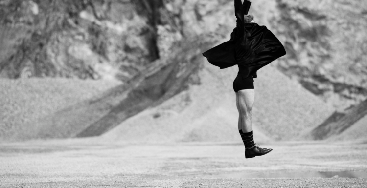 Bilden är i svartvitt och visar en person som hoppar eller svävar i luften i ett kargt, stenigt landskap – kanske en öken eller ett stenbrott. Personen bär en mörk, fladdrande kappa eller rock, korta byxor, långa strumpor och oxford skor. Kroppen är sträckt uppåt, med armarna höjda och händerna vända mot himlen, vilket ger en känsla av rörelse, frihet och uttrycksfullhet – nästan som ett danssteg eller ett konstnärligt språng. Bakgrunden är oskarp, vilket framhäver personen som huvudmotiv och förstärker känslan av dramatik och kontrast.
