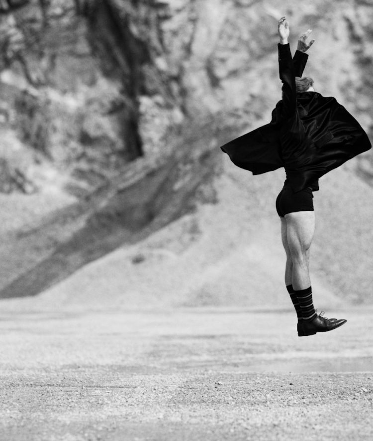 Bilden är i svartvitt och visar en person som hoppar eller svävar i luften i ett kargt, stenigt landskap – kanske en öken eller ett stenbrott. Personen bär en mörk, fladdrande kappa eller rock, korta byxor, långa strumpor och oxford skor. Kroppen är sträckt uppåt, med armarna höjda och händerna vända mot himlen, vilket ger en känsla av rörelse, frihet och uttrycksfullhet – nästan som ett danssteg eller ett konstnärligt språng. Bakgrunden är oskarp, vilket framhäver personen som huvudmotiv och förstärker känslan av dramatik och kontrast.
