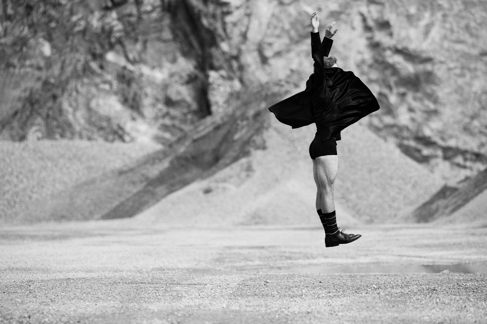 Bilden är i svartvitt och visar en person som hoppar eller svävar i luften i ett kargt, stenigt landskap – kanske en öken eller ett stenbrott. Personen bär en mörk, fladdrande kappa eller rock, korta byxor, långa strumpor och oxford skor. Kroppen är sträckt uppåt, med armarna höjda och händerna vända mot himlen, vilket ger en känsla av rörelse, frihet och uttrycksfullhet – nästan som ett danssteg eller ett konstnärligt språng. Bakgrunden är oskarp, vilket framhäver personen som huvudmotiv och förstärker känslan av dramatik och kontrast.