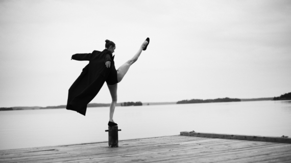 Bilden är i svartvitt och visar en person som står på en brygga vid vattnet. Personen balanserar elegant på en stolpe med ena benet högt uppsträckt i luften, vilket ger en känsla av rörelse och balans. De bär en lång, mörk rock som fladdrar något i luften, samt skor. Bakgrunden är stillsam med en spegelblank sjö och några öar eller landkonturer i fjärran under en molnig himmel. Bilden förmedlar en känsla av frihet, styrka och konstnärlig uttrycksfullhet, med en tydlig kontrast mellan den dynamiska posen och det lugna landskapet.