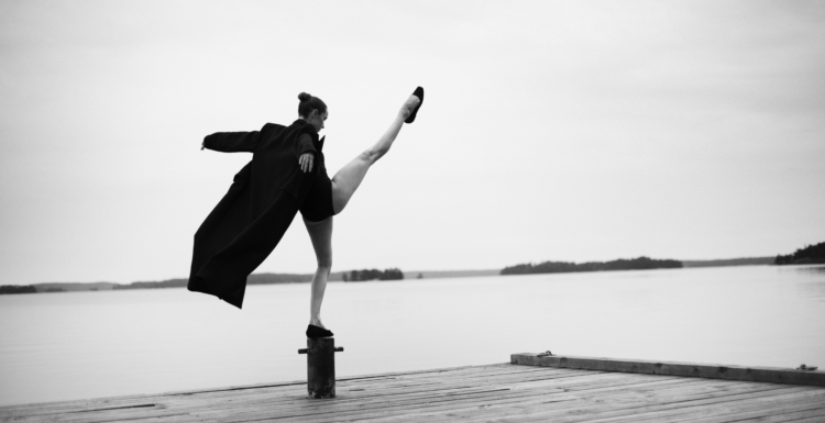 Bilden är i svartvitt och visar en person som står på en brygga vid vattnet. Personen balanserar elegant på en stolpe med ena benet högt uppsträckt i luften, vilket ger en känsla av rörelse och balans. De bär en lång, mörk rock som fladdrar något i luften, samt skor. Bakgrunden är stillsam med en spegelblank sjö och några öar eller landkonturer i fjärran under en molnig himmel. Bilden förmedlar en känsla av frihet, styrka och konstnärlig uttrycksfullhet, med en tydlig kontrast mellan den dynamiska posen och det lugna landskapet.