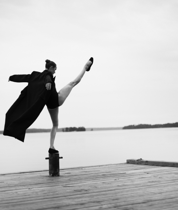 Bilden är i svartvitt och visar en person som står på en brygga vid vattnet. Personen balanserar elegant på en stolpe med ena benet högt uppsträckt i luften, vilket ger en känsla av rörelse och balans. De bär en lång, mörk rock som fladdrar något i luften, samt skor. Bakgrunden är stillsam med en spegelblank sjö och några öar eller landkonturer i fjärran under en molnig himmel. Bilden förmedlar en känsla av frihet, styrka och konstnärlig uttrycksfullhet, med en tydlig kontrast mellan den dynamiska posen och det lugna landskapet.
